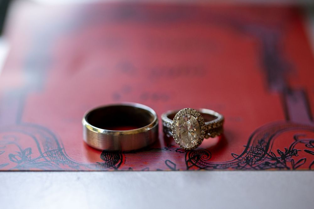 Wedding rings on a red ornate invitation, featuring a brushed metal men's band beside a diamond engagement ring with a pavé halo setting.