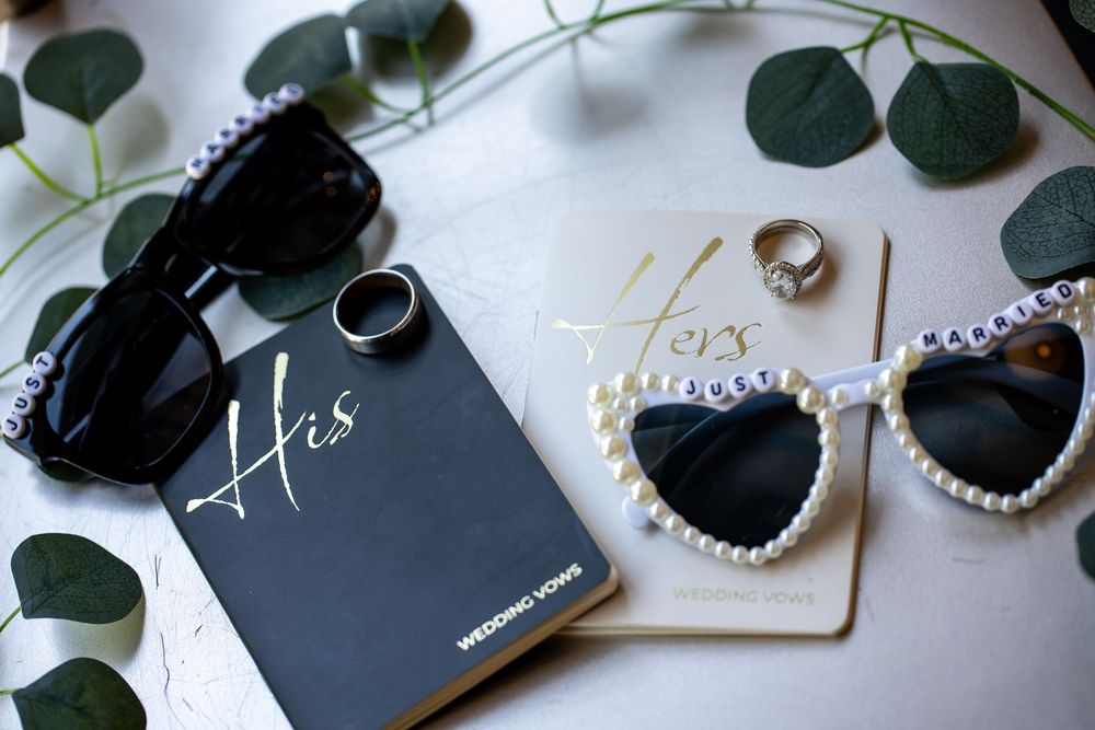 His and Hers wedding vow books with black sunglasses, pearl heart-shaped sunglasses and wedding rings styled among eucalyptus leaves.