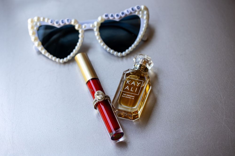 Heart-shaped pearl-trimmed sunglasses, a red lip gloss with gold cap, and a Kayali perfume bottle arranged on a gray surface.