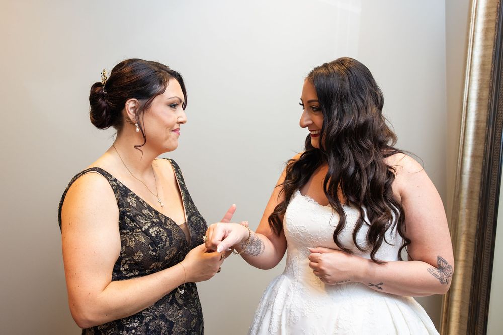 Bride in a white strapless wedding gown with long wavy hair shares a tender moment with a woman in a black lace dress holding her hands.