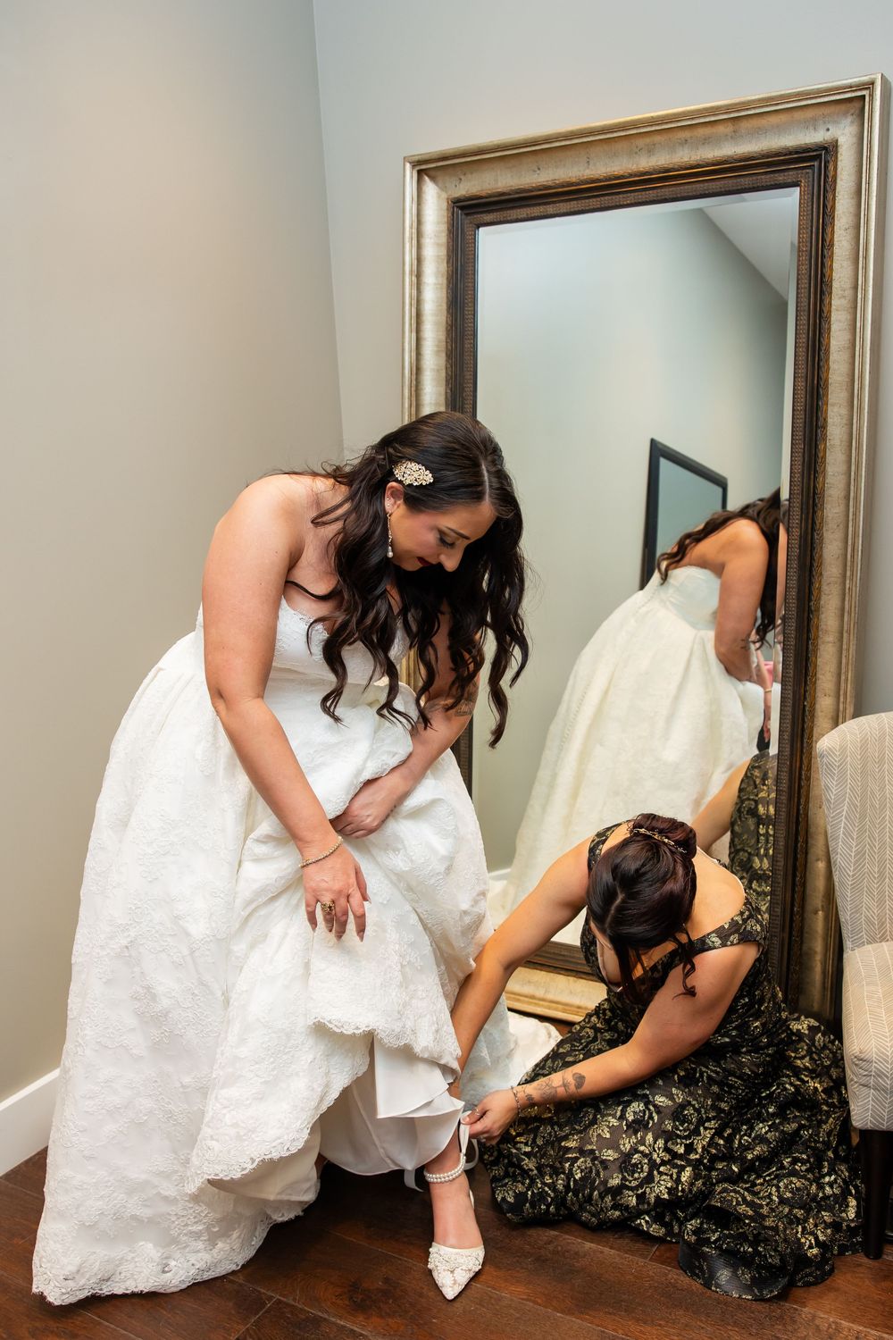 Bride in a white lace wedding gown stands before a gold-framed mirror while a bridesmaid in a green dress kneels to adjust her shoe.