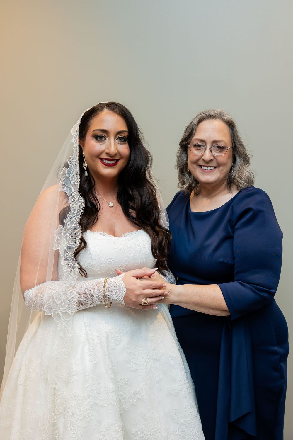 A smiling bride in a lace wedding gown and veil holds hands with an older woman in a navy blue dress on the wedding day.