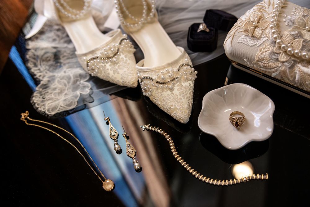 Lace embellished bridal heels, pearl bracelet, drop earrings, pendant necklace and ring dish arranged on a reflective black surface.