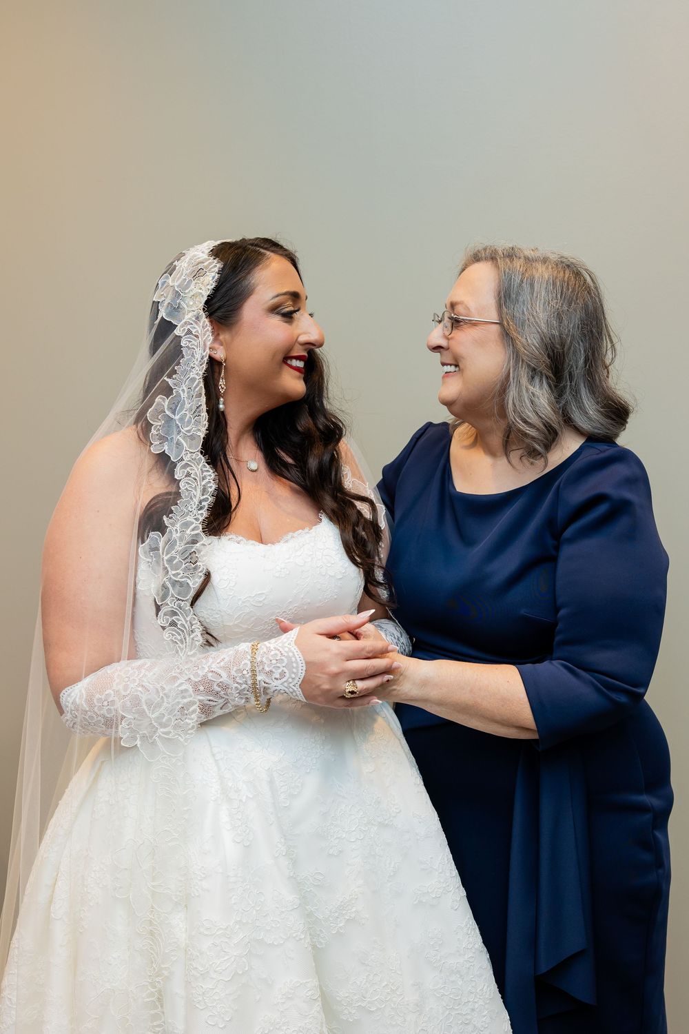 Smiling bride in a white lace gown and veil holds hands with an older woman in a navy dress in a tender wedding day moment.