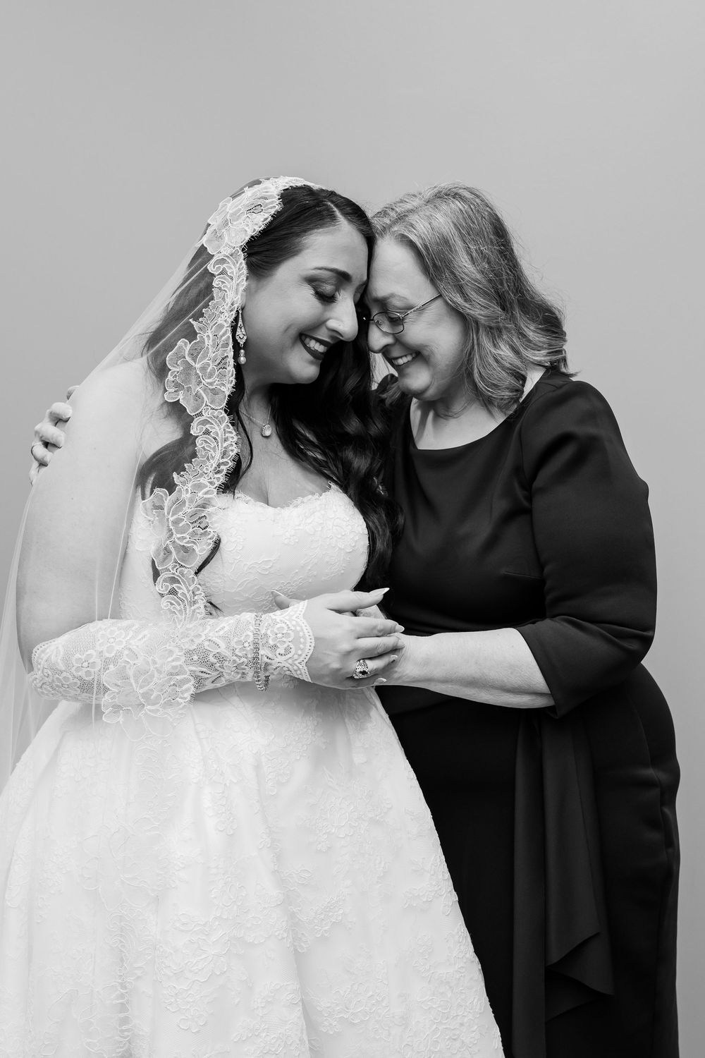 Black and white portrait of a smiling bride in a lace veil and gown touching foreheads with an older woman in a dark dress, holding hands.