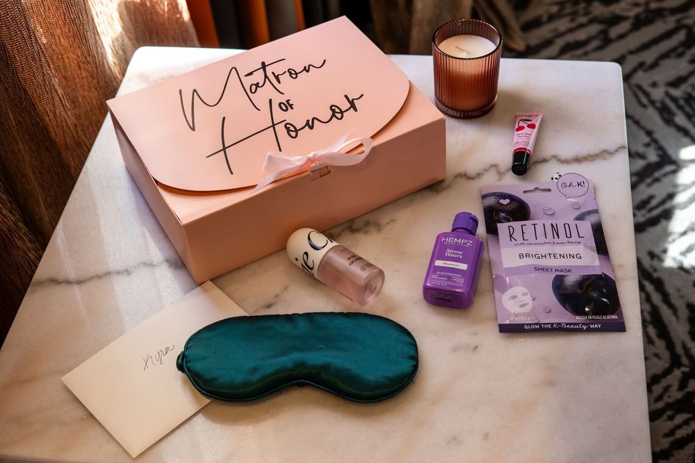 Pink Matron of Honor gift box with teal silk sleep mask, retinol face mask, purple lotion, lip gloss, candle and card on a marble table.