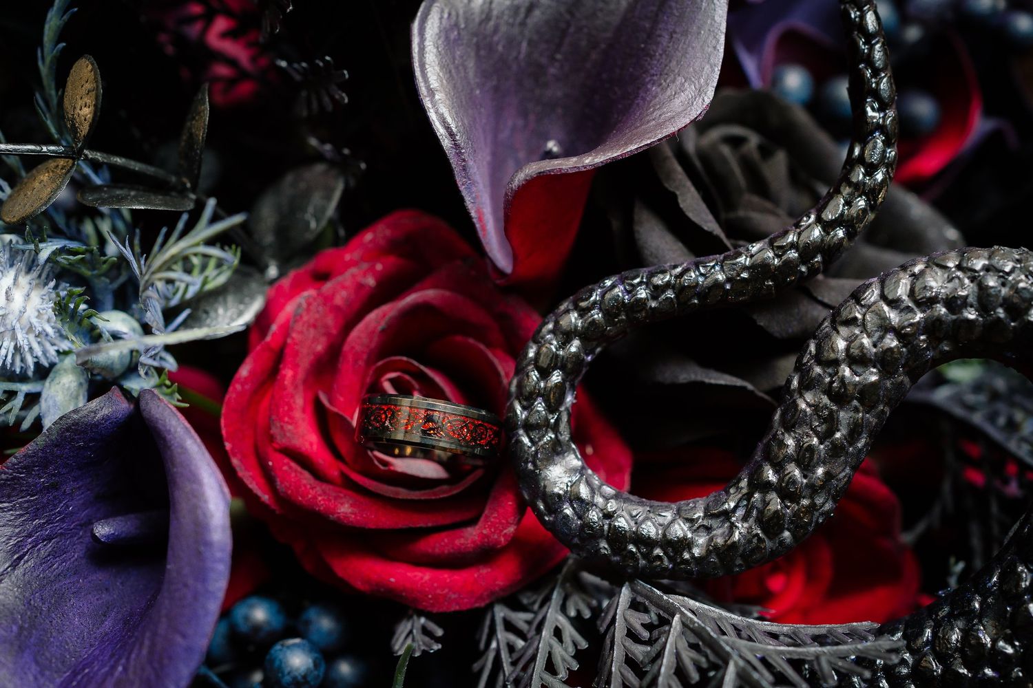 Gothic wedding ring resting on a red rose, surrounded by purple calla lilies, dark blooms, and a coiled black snake figurine.