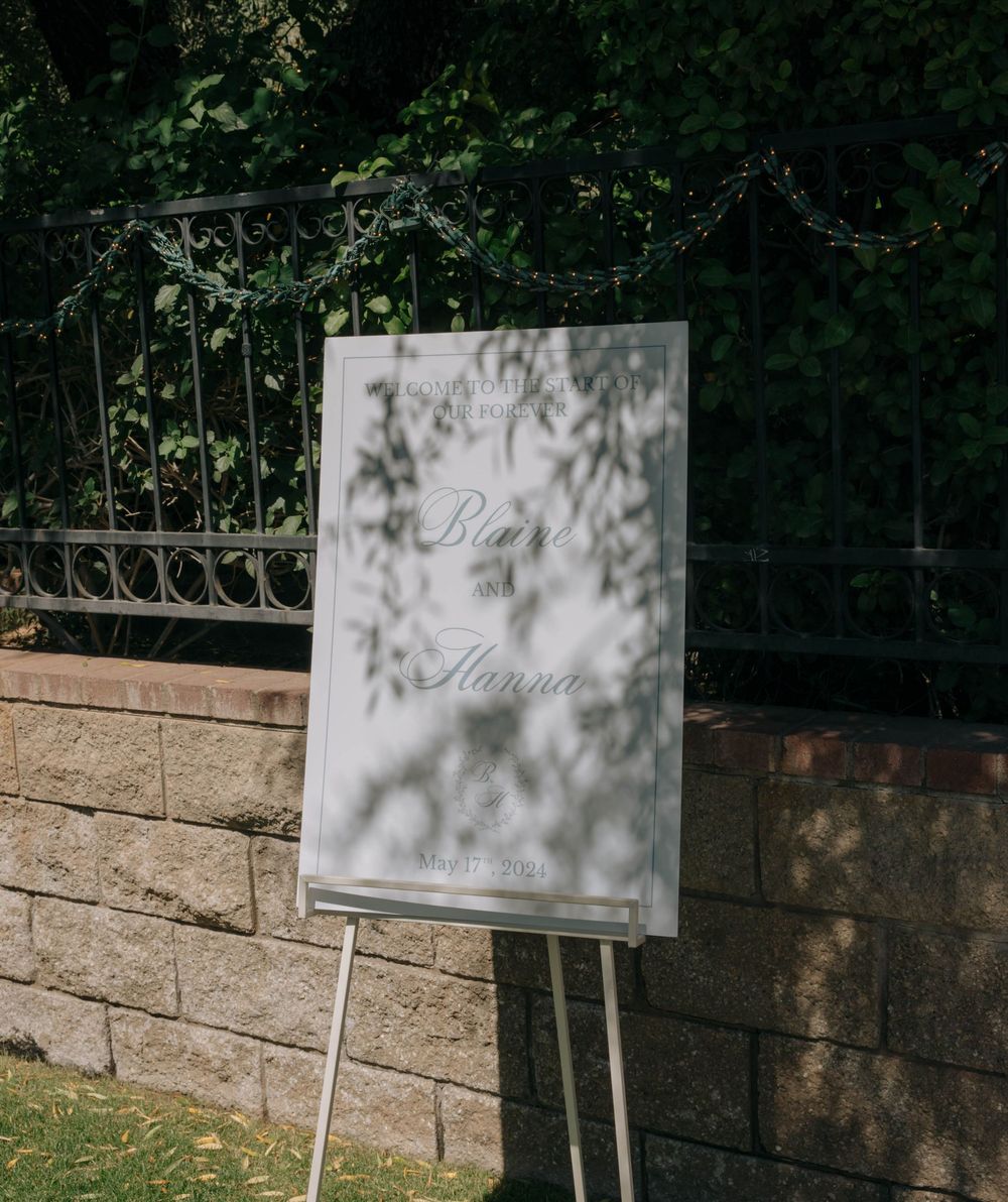 Elegant wedding welcome sign with names in calligraphy on an easel before a wrought iron fence and leafy dappled shade.