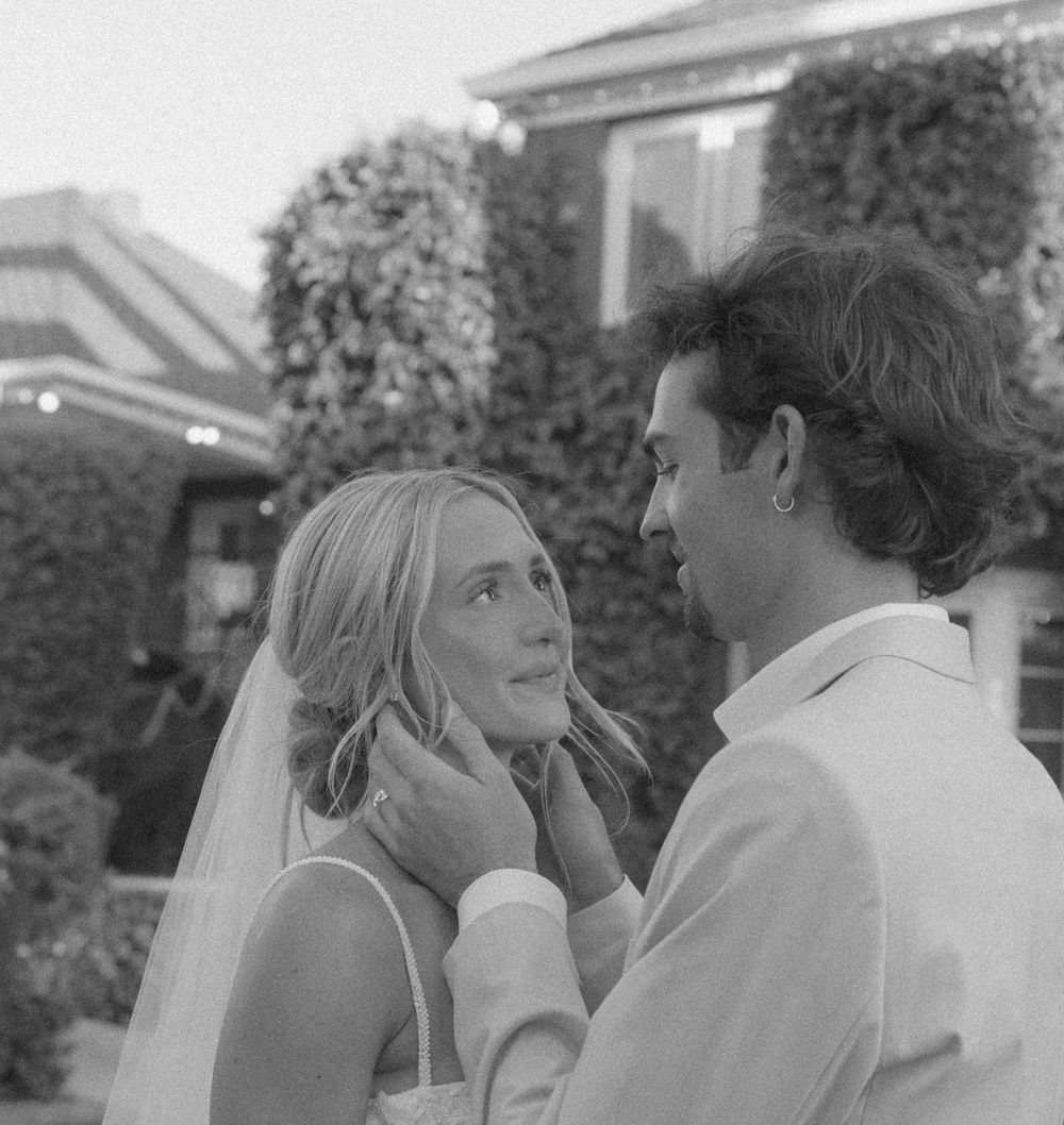 Black and white portrait of a groom gently holding his veiled bride's face as she gazes up at him outdoors.