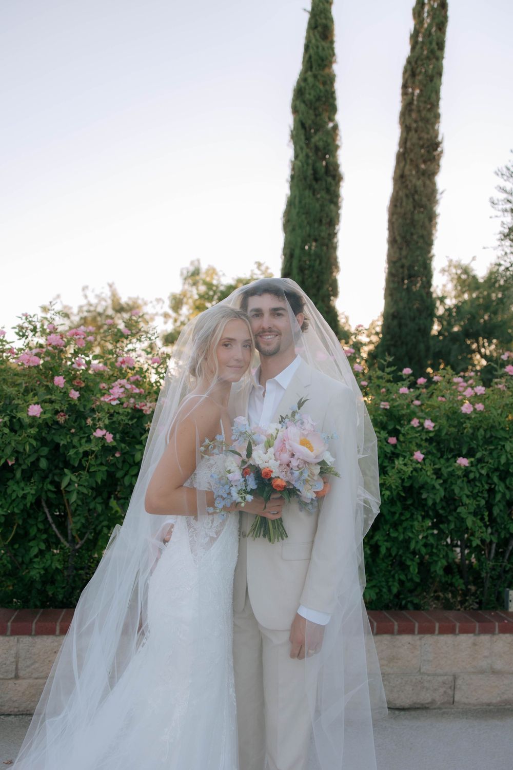 Bride and groom embrace under a sheer veil holding a pastel bouquet, with tall cypress trees and pink flowering bushes behind them.