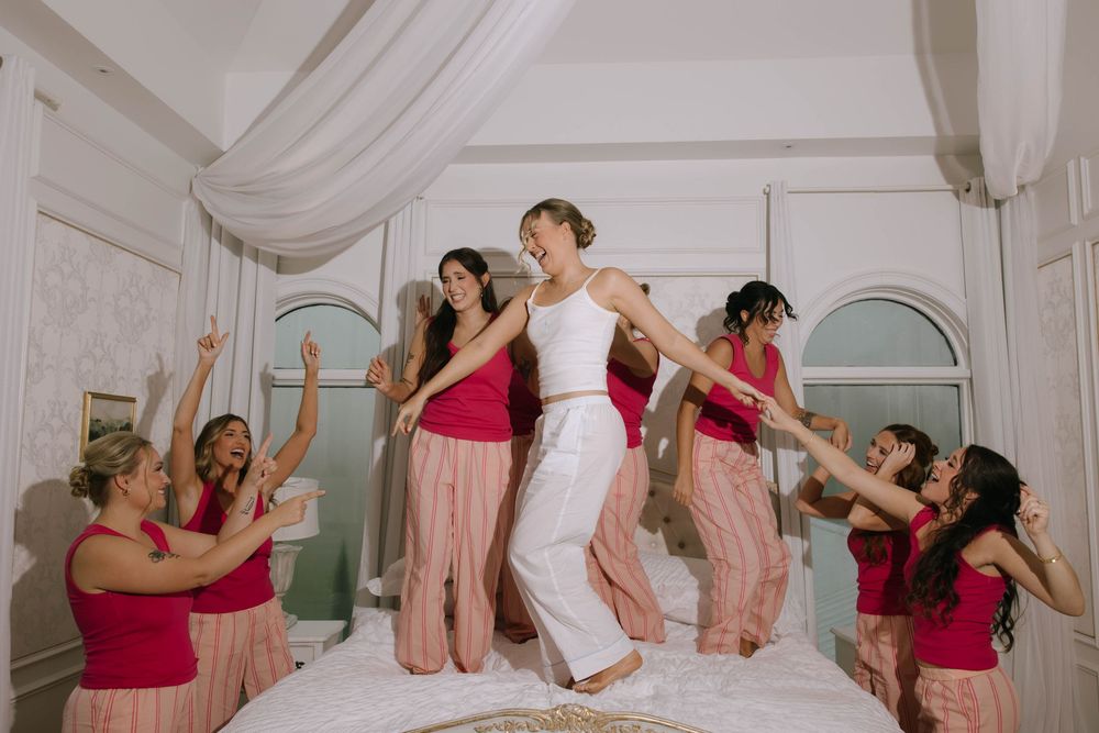 A bride and her bridesmaids in pink tops and matching pants dance and cheer together on a white tufted bed.