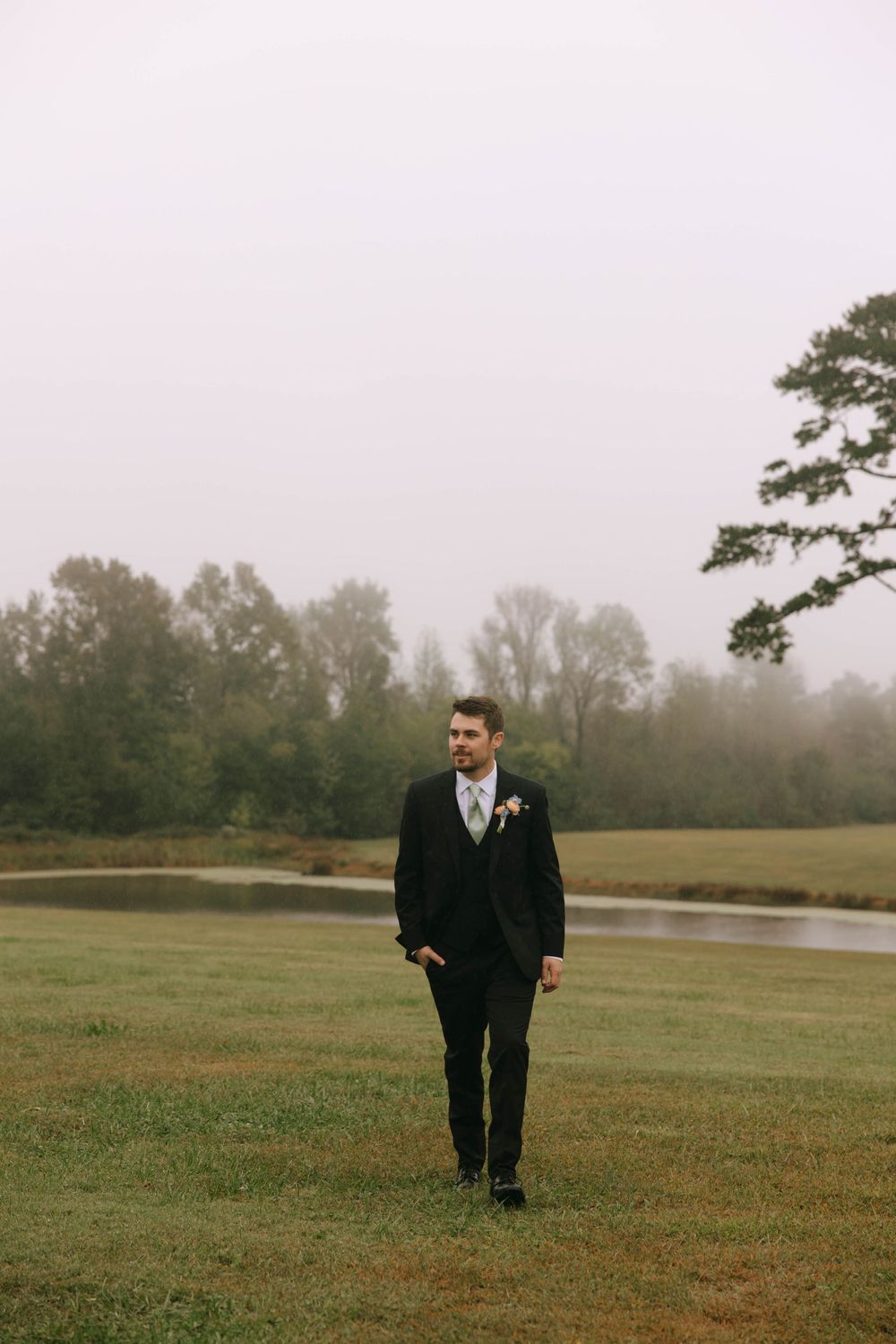 Groom in a dark suit with a boutonniere walks across a misty green lawn beside a pond and tall trees.