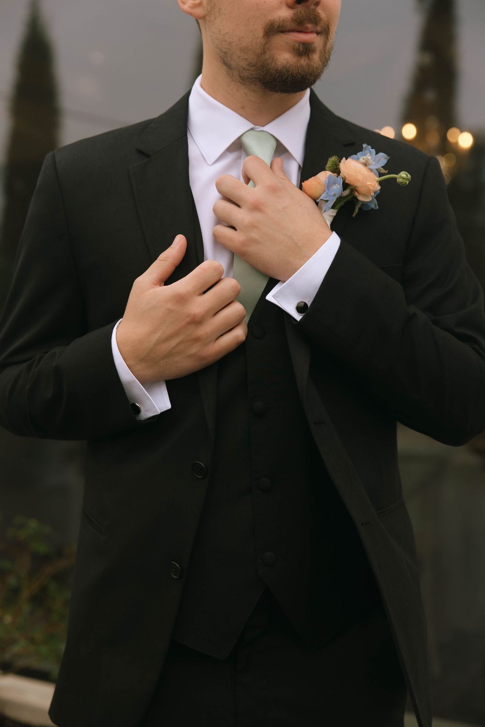 Groom in a black three-piece suit and sage green tie adjusts his collar, wearing a peach and blue floral boutonniere.