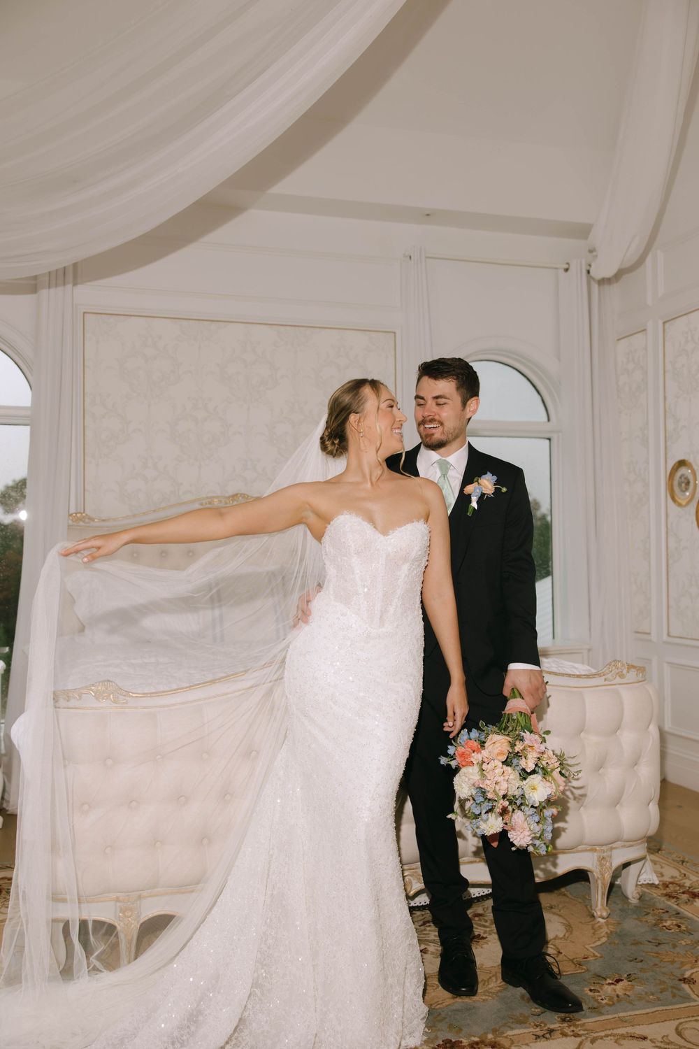 Bride in a sparkling fitted wedding gown and long veil holds a bouquet beside her smiling groom in an elegant white room.