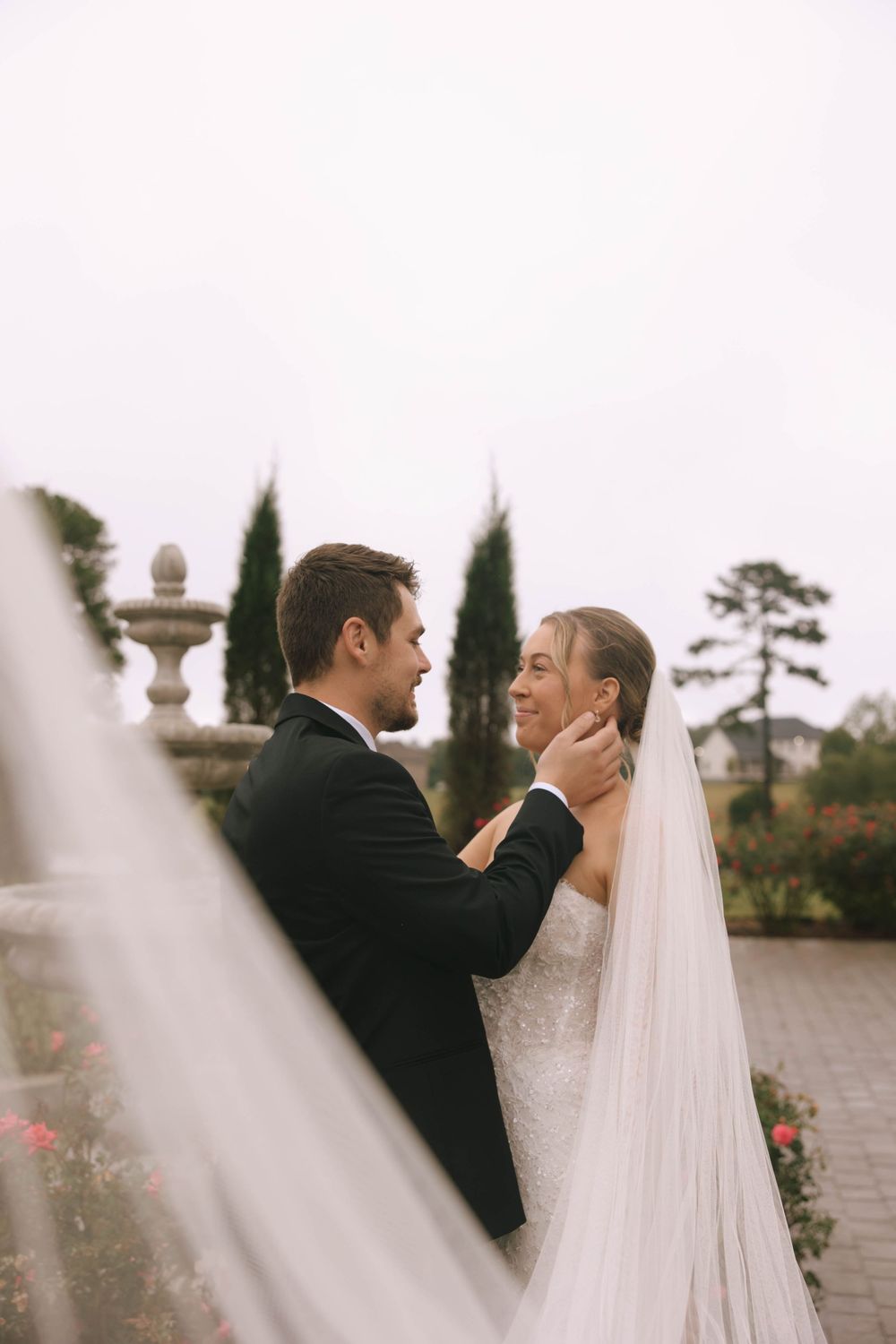 Groom gently cradles his smiling bride's face in a garden with a stone fountain, her long veil flowing in the foreground.