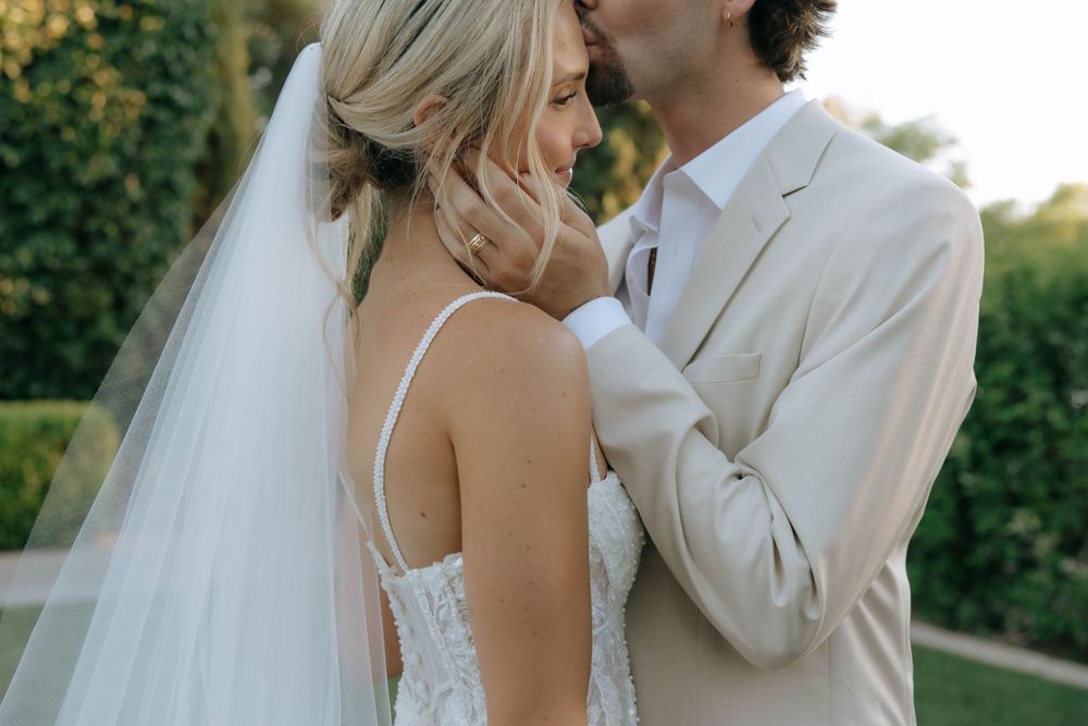 Groom gently touches the bride's cheek as they share an intimate moment, her long veil flowing in a sunlit garden.