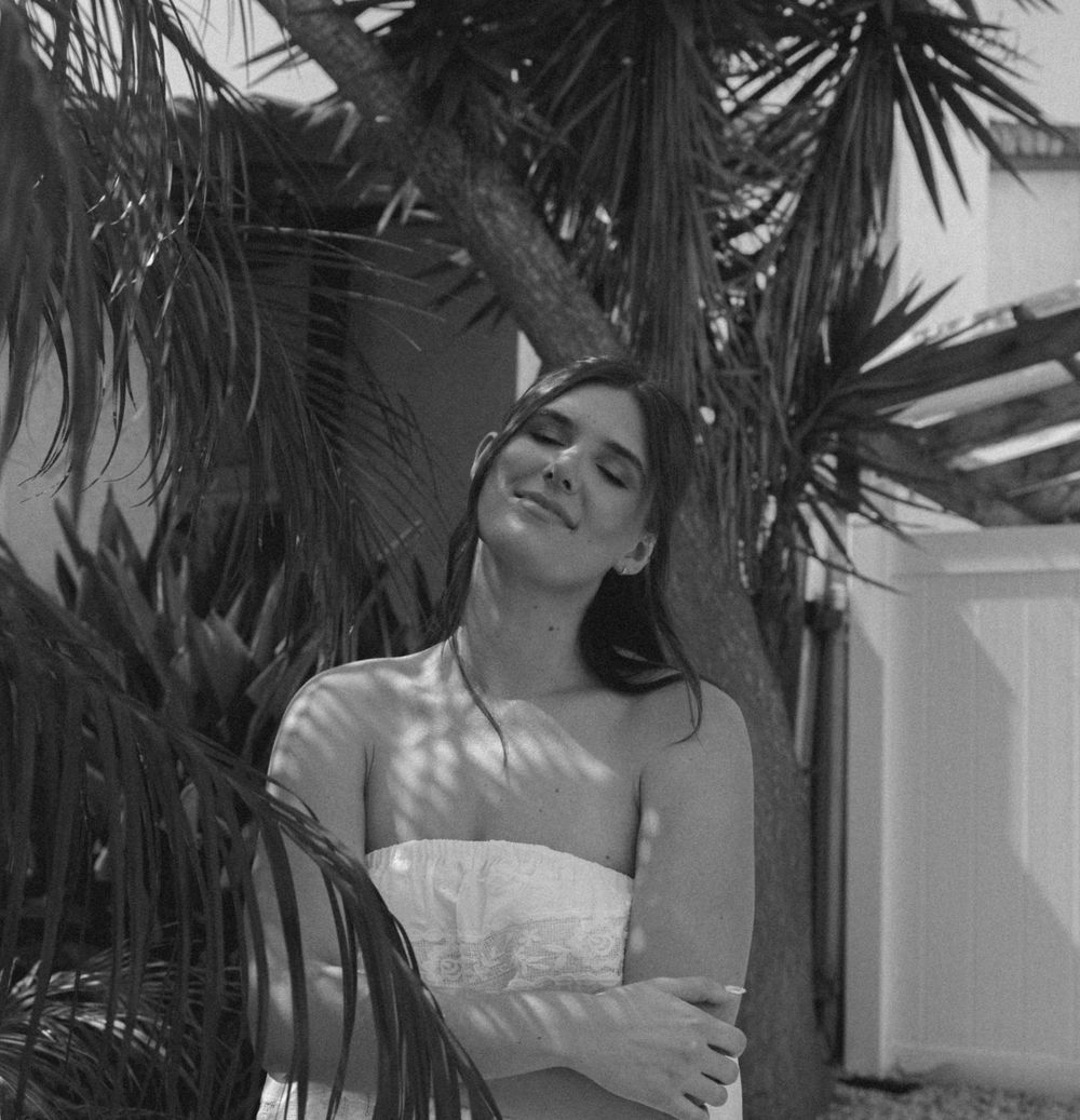 Black and white portrait of a smiling bride in a strapless dress leaning against a palm tree with eyes closed.