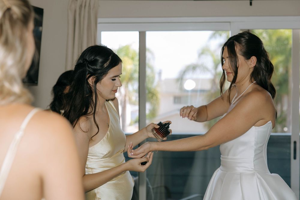 Bridesmaid helps the bride in a white gown with perfume spray while getting ready by a sunny window.