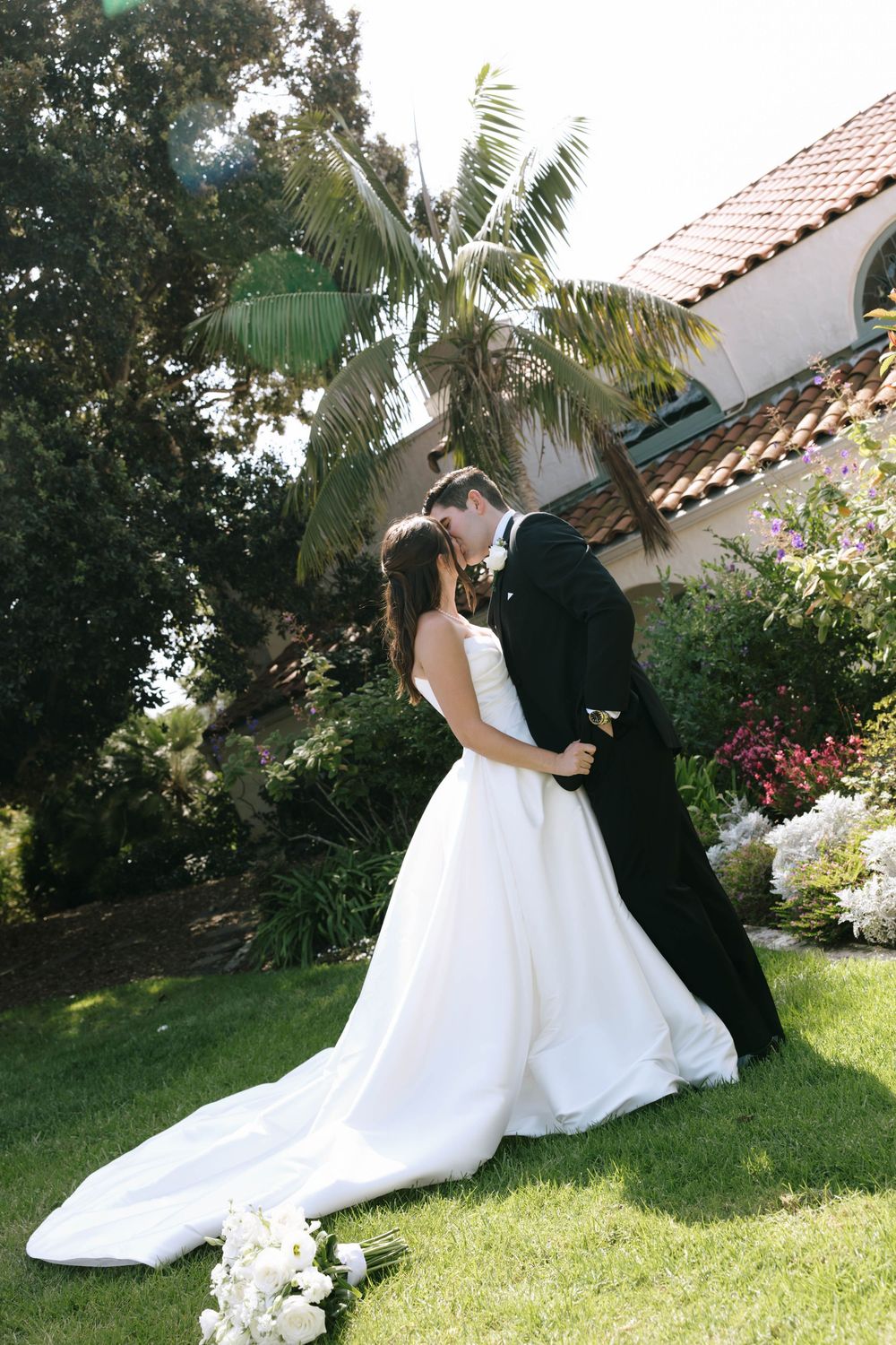 Groom in a black tuxedo kisses his bride in a white gown with a long train on a green lawn near a palm tree.