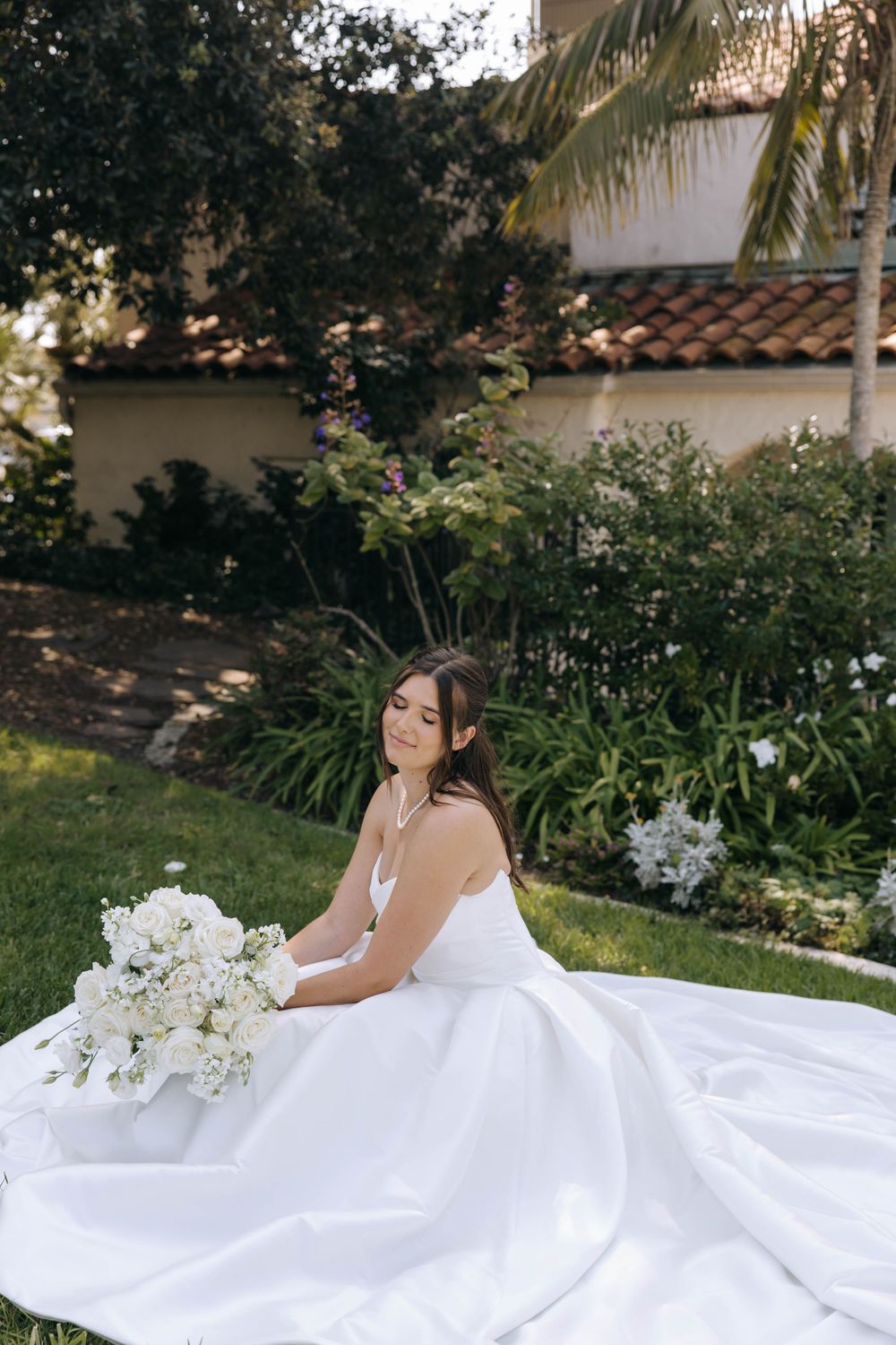 Bride in a strapless white gown sits on the lawn with her eyes closed, holding a white bouquet in a garden with a tile-roofed building.