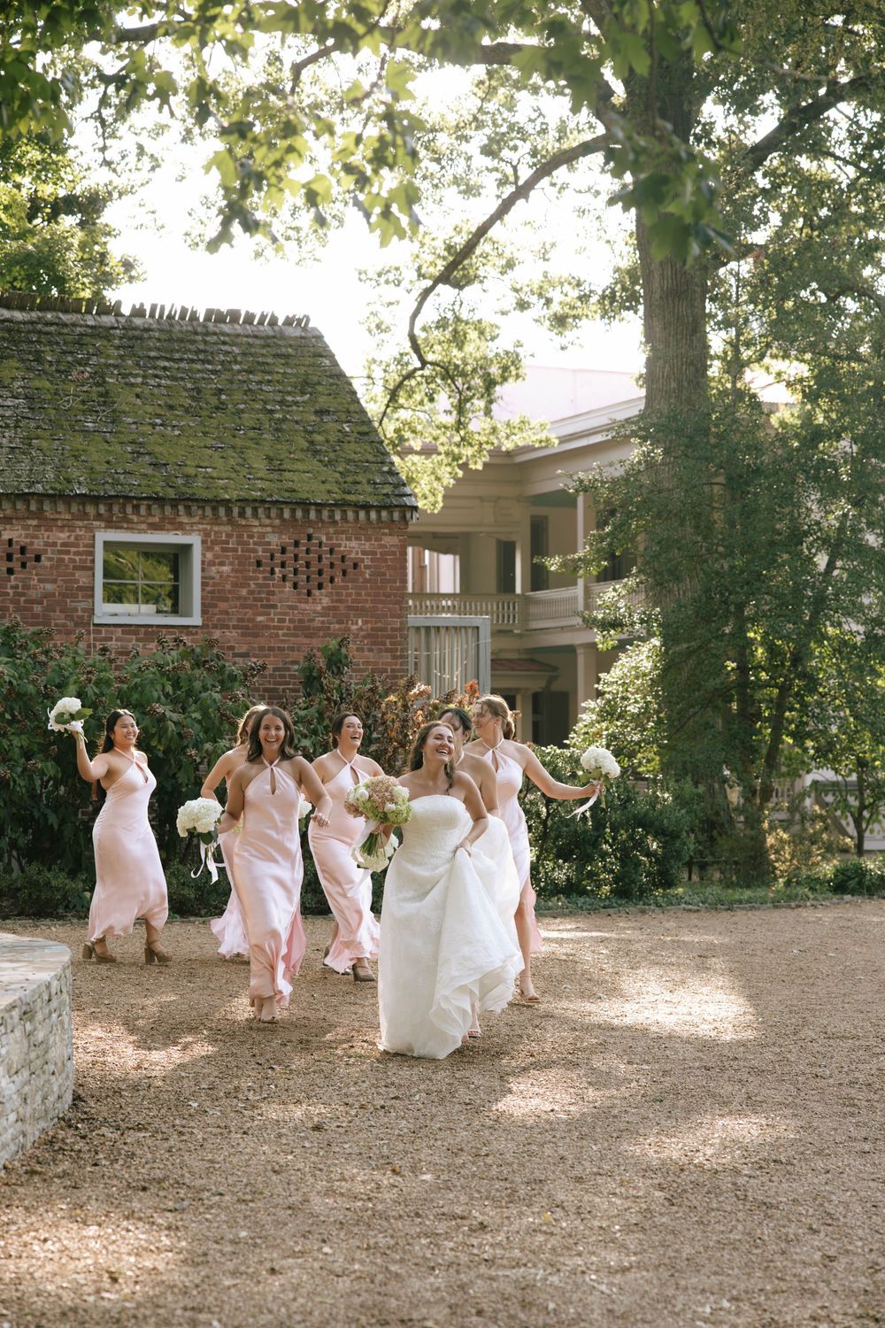 Bride in a white gown and bridesmaids in blush dresses laugh and run together holding bouquets on a garden path.
