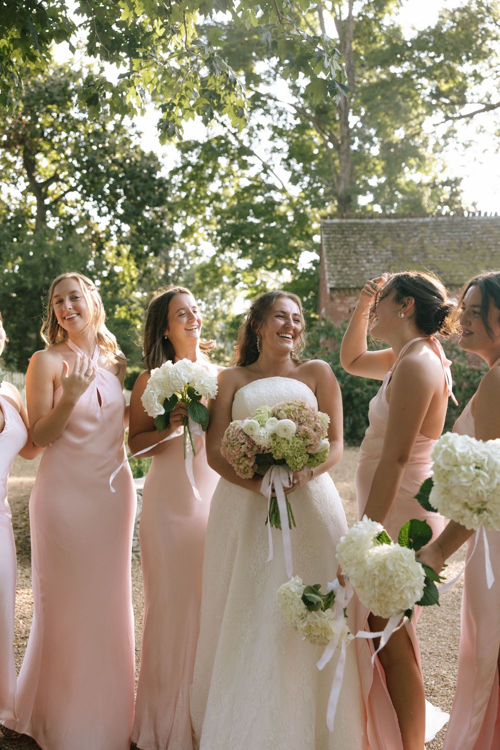 Smiling bride surrounded by bridesmaids in blush satin dresses holding white and pink hydrangea bouquets outdoors.
