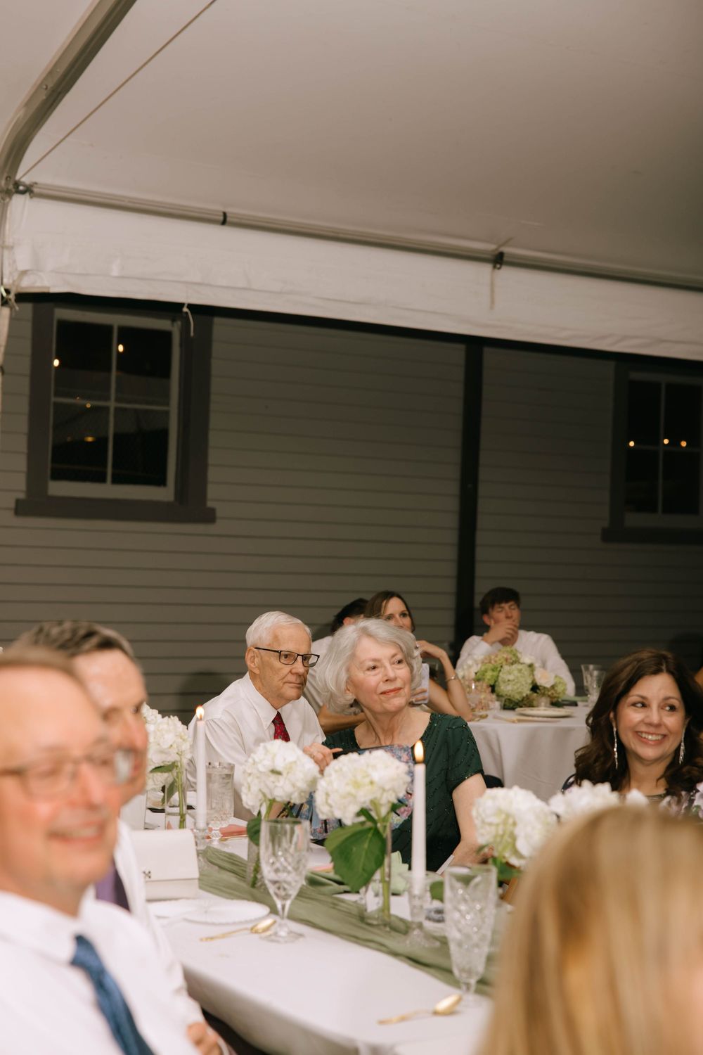 Wedding guests seated at long tables with white hydrangea centerpieces and candles smile during a reception inside a tent.