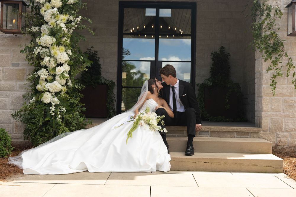Bride and groom share a tender moment on stone steps beside a white floral arrangement, her ballgown spread across the ground.
