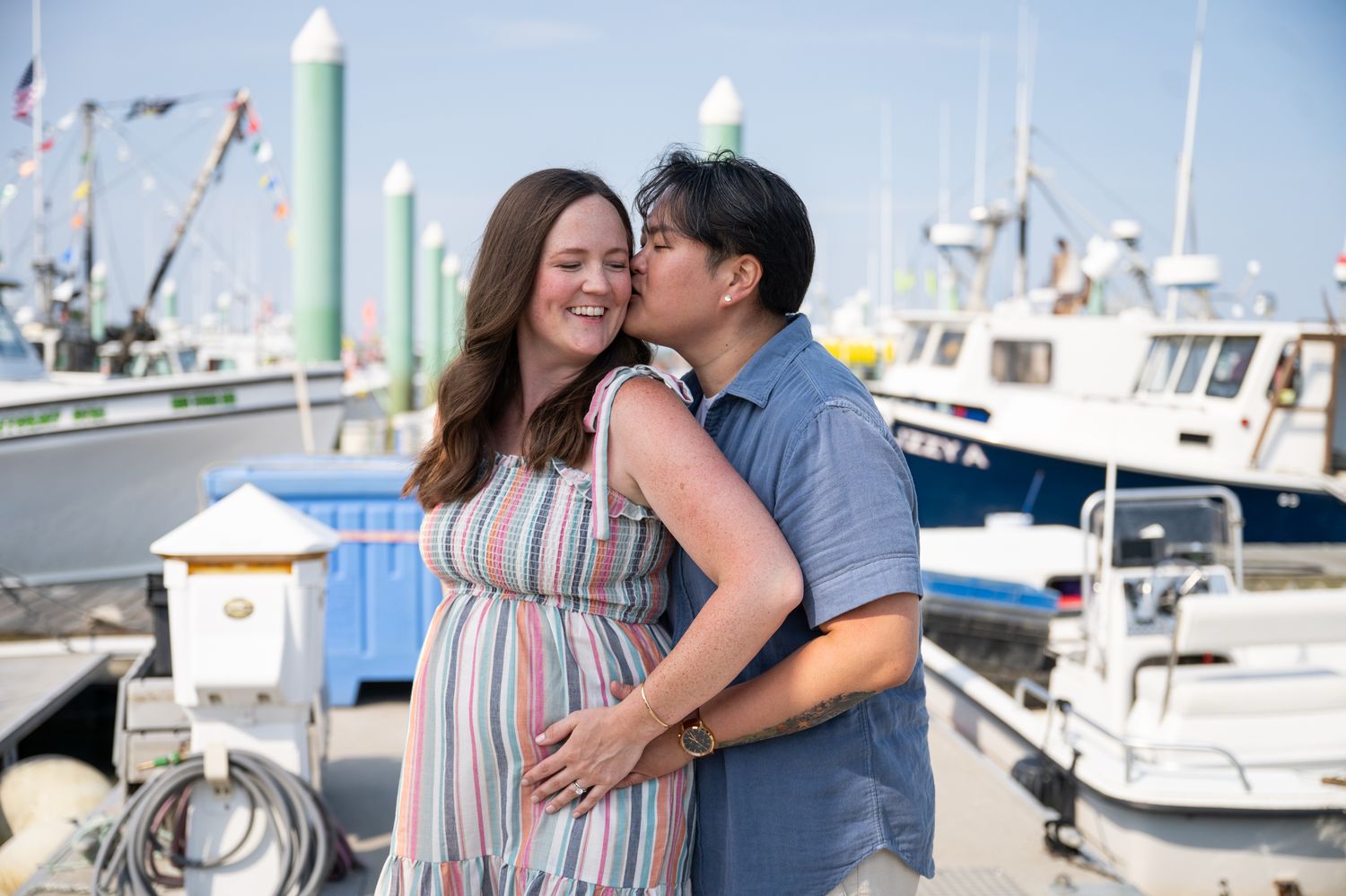 Smiling pregnant woman in a striped dress is kissed on the cheek by her partner during a maternity photo session at a sunny marina.