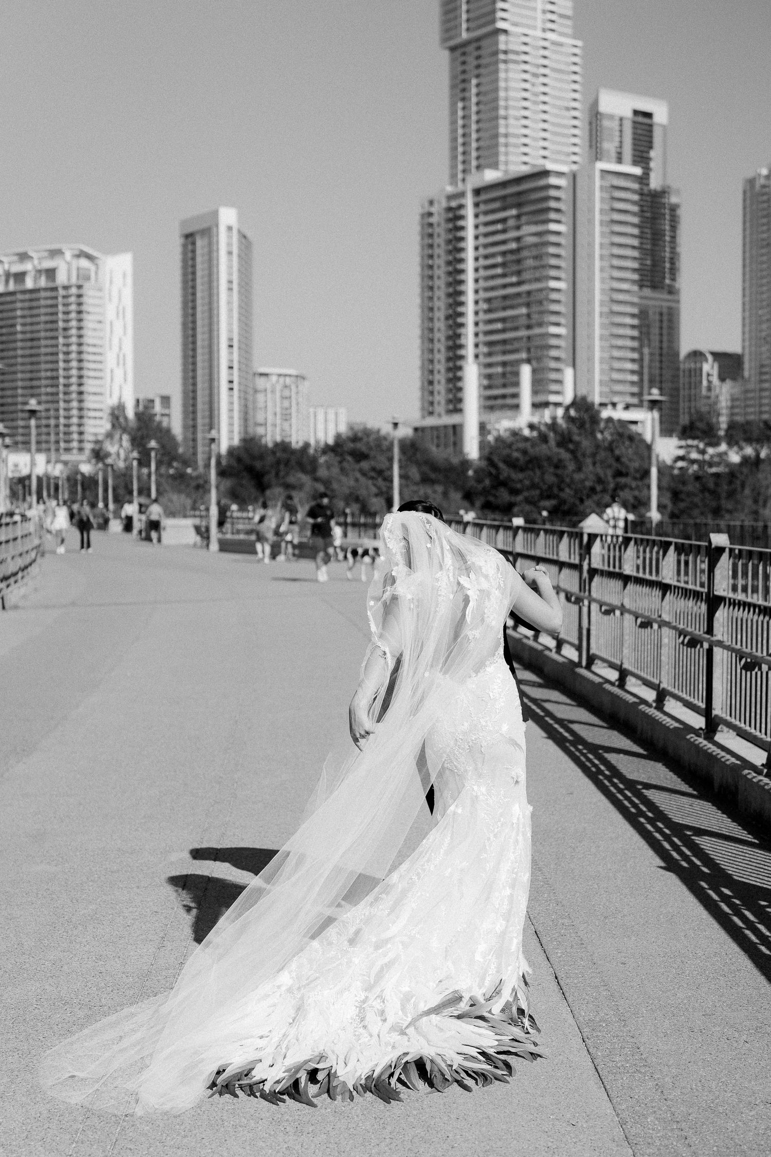Black and white photo of a bride in a flowing gown and veil on a city bridge with skyscrapers in the background.