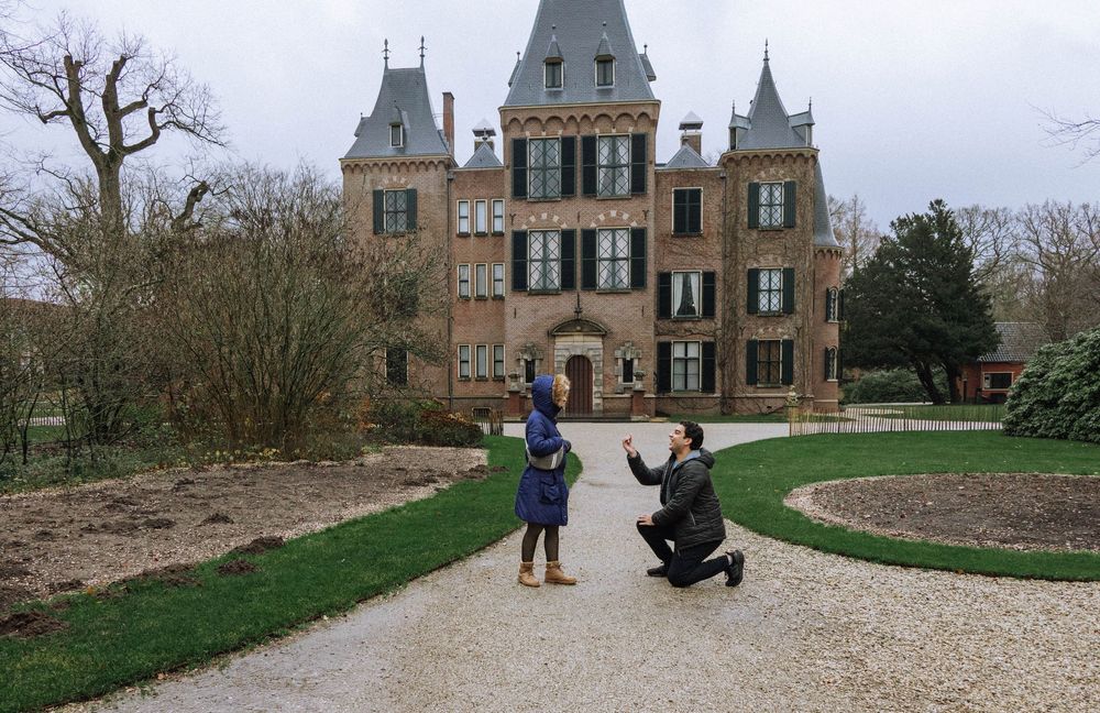 Man proposes to woman on one knee in front of a grand historic castle surrounded by lush gardens. Kokenhof Kasteel