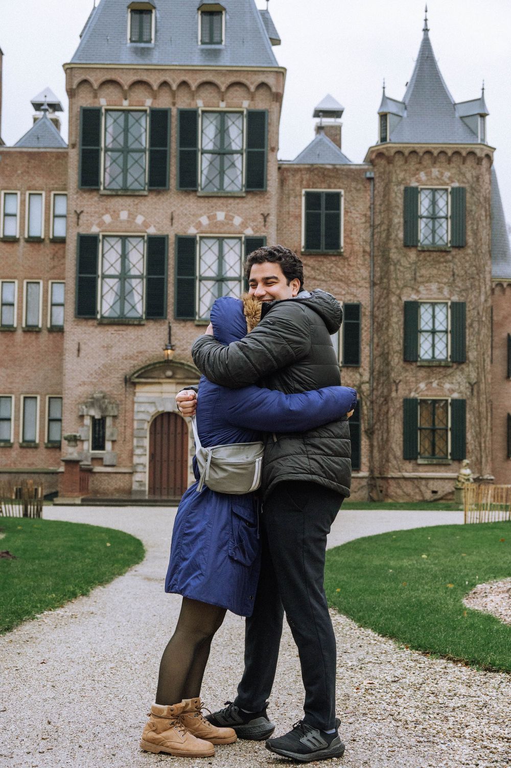 Couple hugging in front of a historic brick castle on a gravel path, wearing winter coats outdoors.