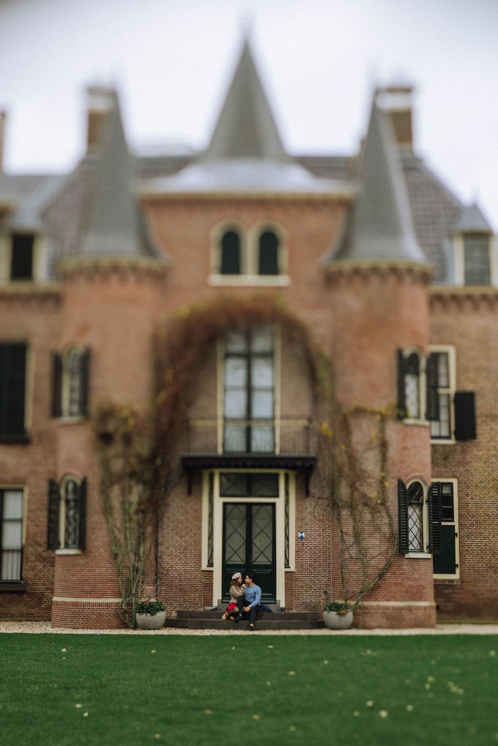 Couple sitting on steps of a grand brick castle with ivy-covered walls, pointed turrets, and lush green lawn. Koukenhof Kasteel