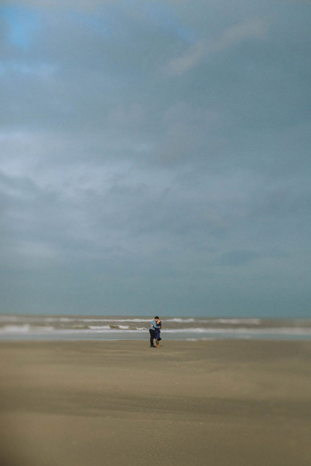A lone person stands on a vast sandy beach under a dramatic cloudy sky with ocean waves in the background.