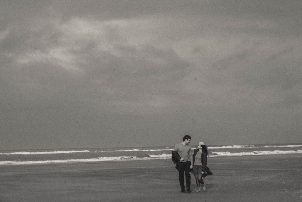 Black and white photo of people standing on a beach with cloudy skies and ocean waves in the background.