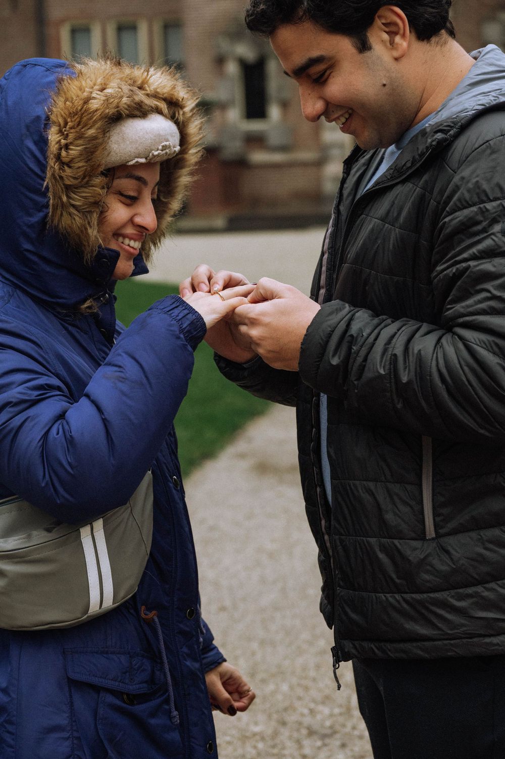A smiling couple in winter jackets sharing a romantic moment as the man places a ring on the woman's finger outdoors.