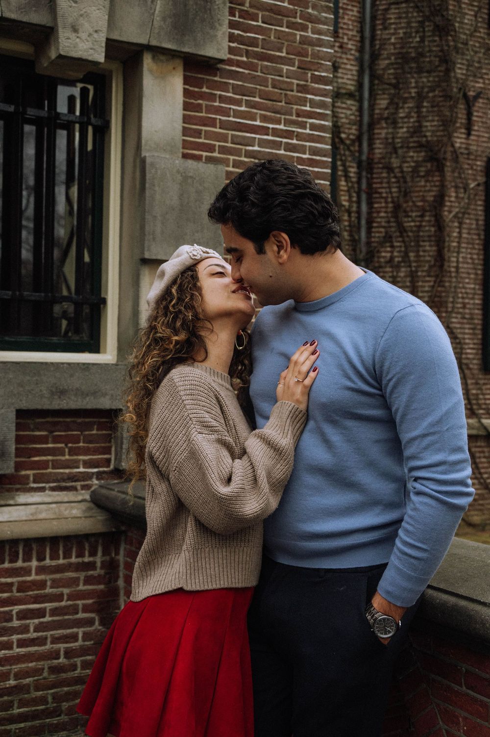A couple shares a romantic kiss outdoors against a brick wall, she wears a red skirt and beige sweater.