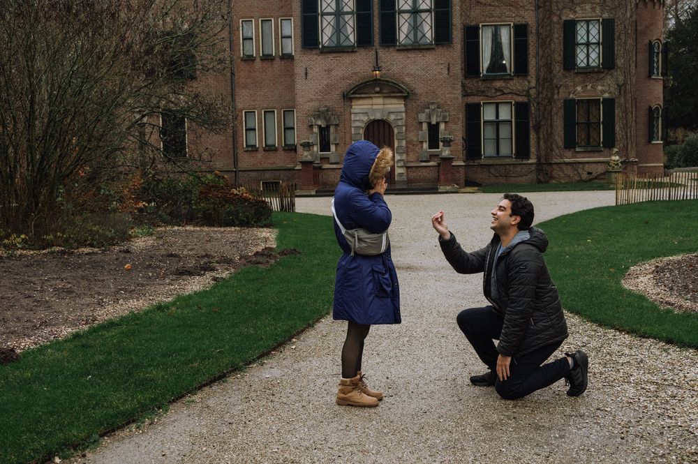 Man proposing on one knee to woman in blue coat outside historic brick mansion in garden setting.