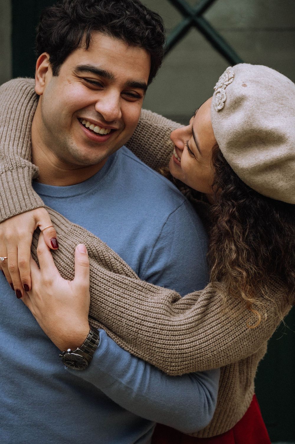 A joyful couple embracing and smiling at each other, woman in beret and tan sweater hugging man in blue shirt outdoors.