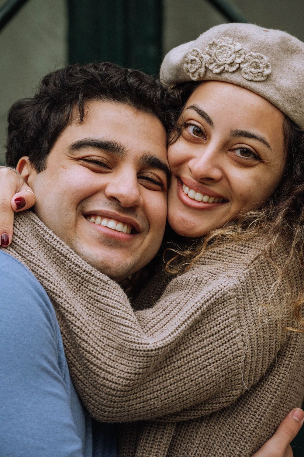 A smiling couple embraces warmly, the woman wearing a beige beret and knit sweater, cheek-to-cheek outdoors.