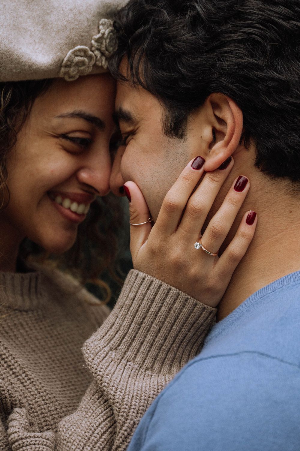 Happy couple sharing a romantic moment, woman wearing engagement ring with dark red nails and beige knit sweater.