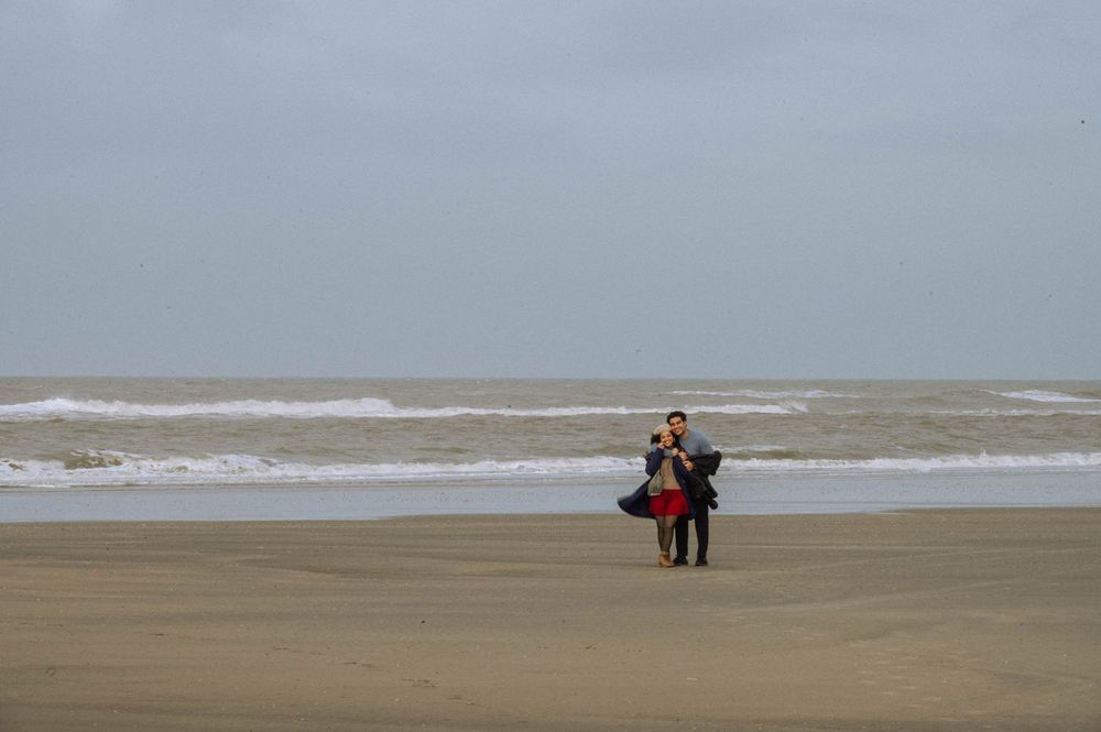 Two people standing on a vast sandy beach with rough ocean waves under an overcast sky.