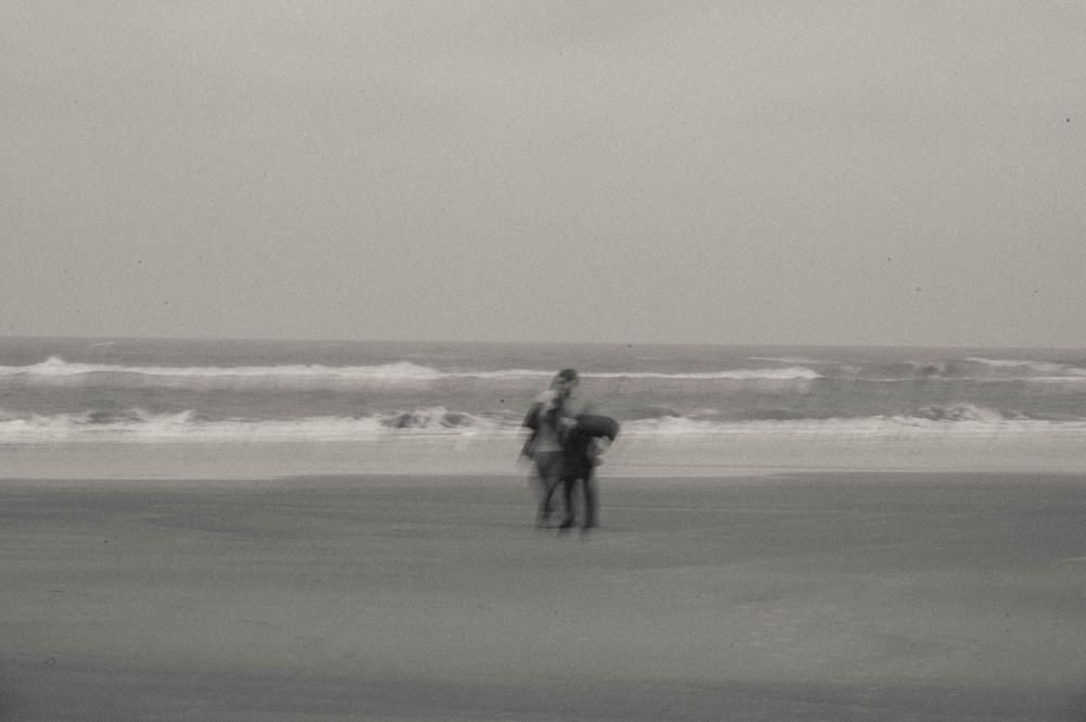 Two people stand on a misty beach with ocean waves in the background, captured in black and white photography.
