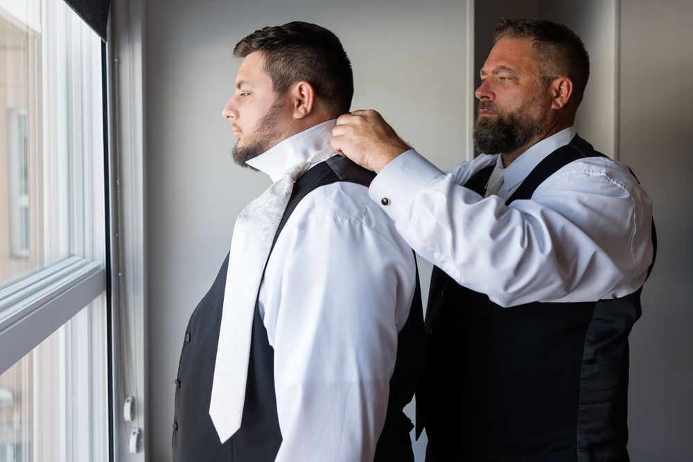 Groomsman in a dark vest helps adjust a white tie on a groom in a white dress shirt while getting ready by a window before the wedding.