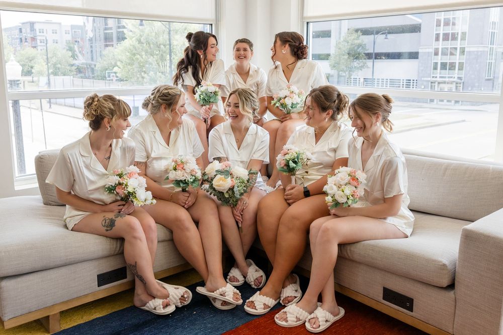 Bride and bridesmaids in white robes and slippers sit on couches holding bouquets of white flowers in a bright room before the wedding.