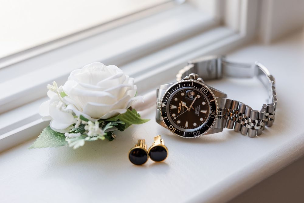 Wedding boutonniere with white rose, silver dive watch and black and gold cufflinks resting on a sunlit windowsill.
