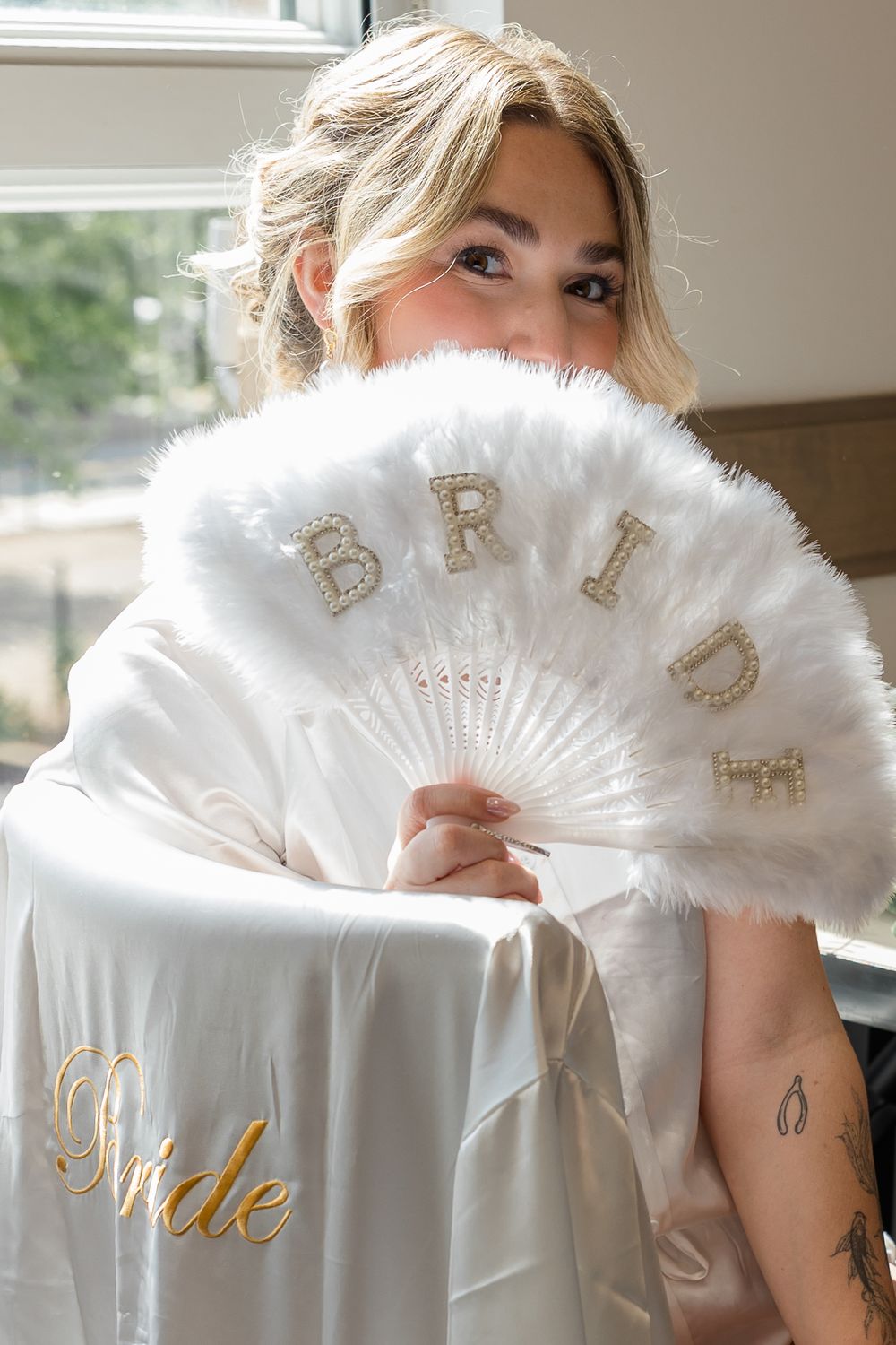 Bride in a white satin robe smiling behind a fluffy white feather fan decorated with gold BRIDE lettering.