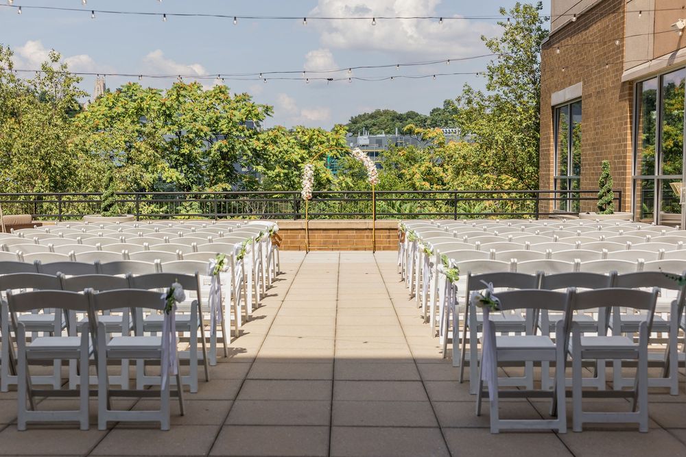 Outdoor wedding ceremony venue with white folding chairs lining a stone aisle toward a wooden altar, surrounded by lush green trees.