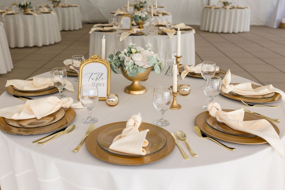 Elegant wedding reception table with gold chargers, folded cream napkins, crystal glasses, white floral centerpiece, candlesticks and table 
