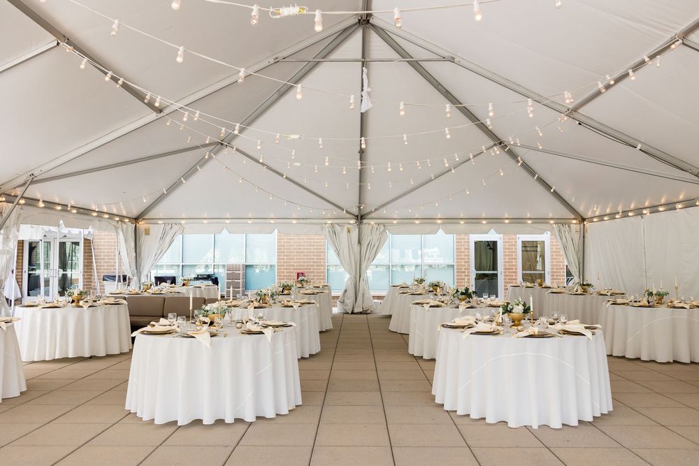 Elegant white wedding reception tent with round tables, linens, floral centerpieces, string lights and stone flooring, ready for guests.